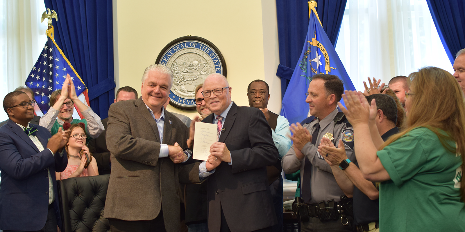 A Crowd of people applaud while two men in the center shake hands, while one displays a signed document