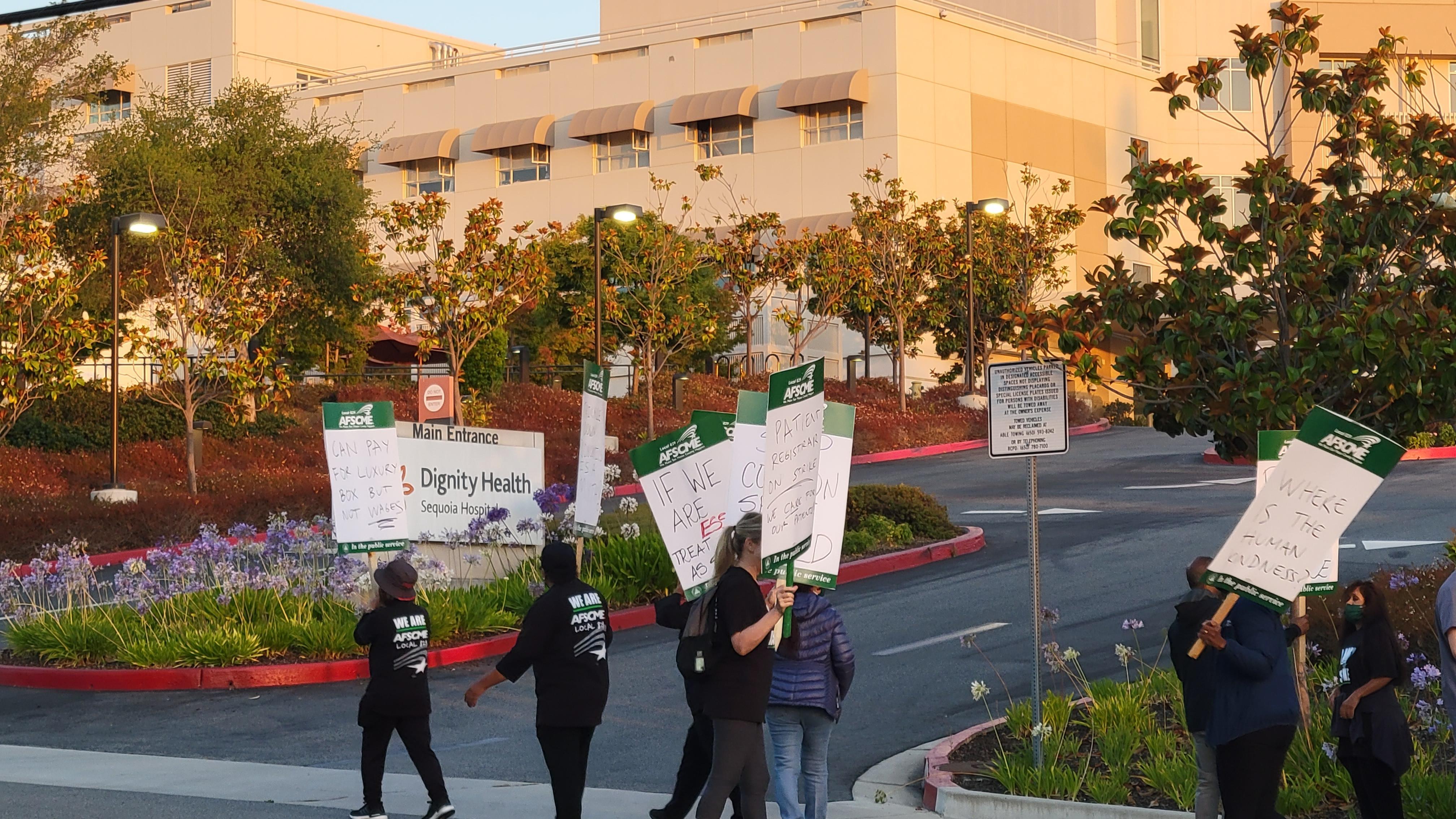 Image of workers holding picket signs and walking past a hospital