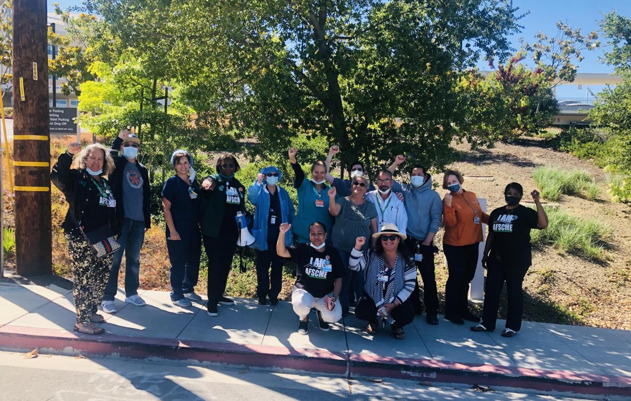 Image of hospital workers standing on sidewalk outside of Sequoia Hospital, raising their fists and wearing AFSCME shirts