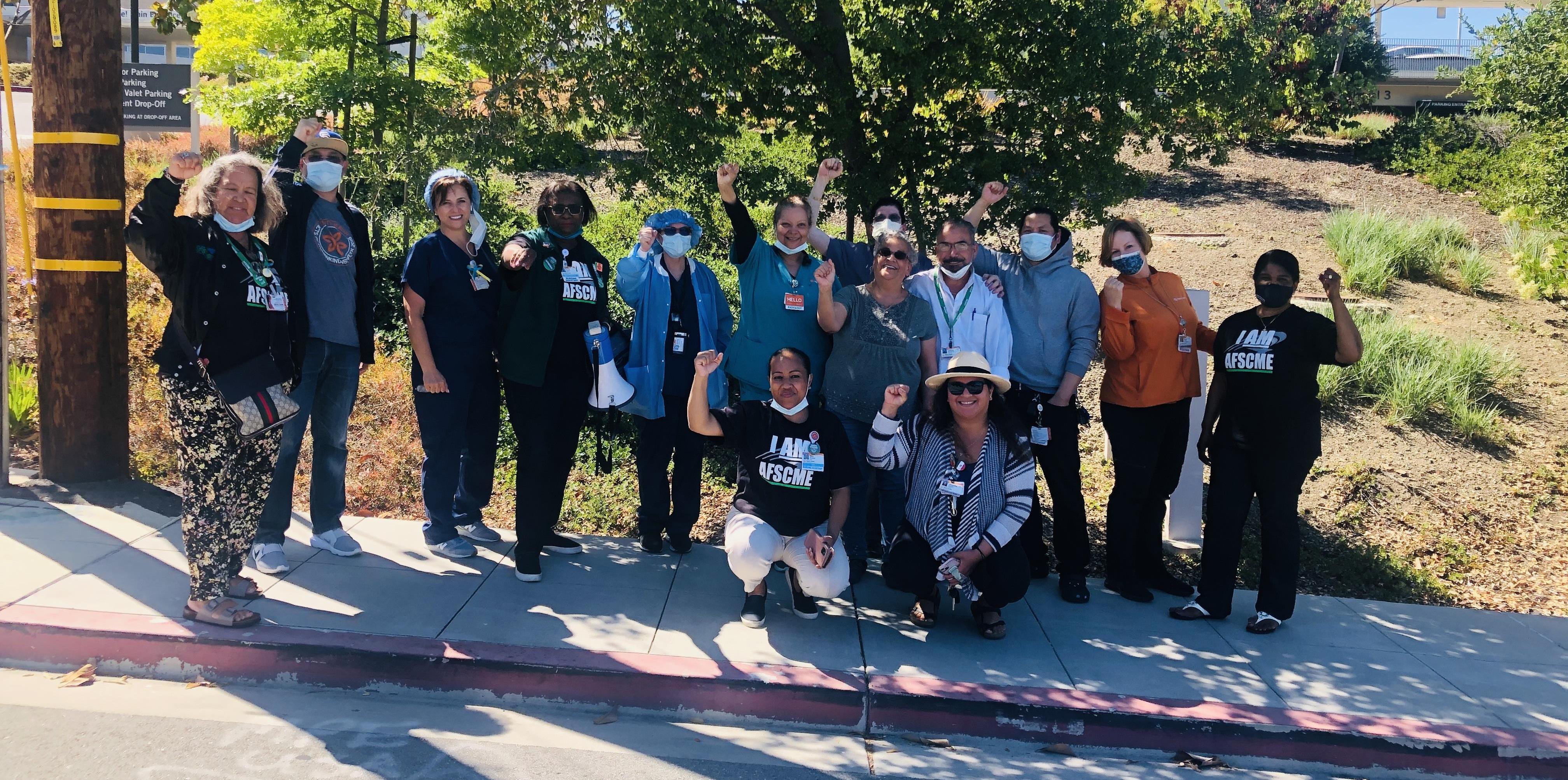 Image of hospital workers standing on sidewalk outside of Sequoia Hospital, raising their fists and wearing AFSCME shirts